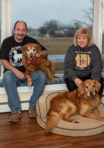 A couple sitting by a window with their two golden retrievers, one on a cushion on the floor and the other sitting between them.