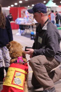 A man kneels beside a service dog in a vest, holding a leash, at an indoor event.