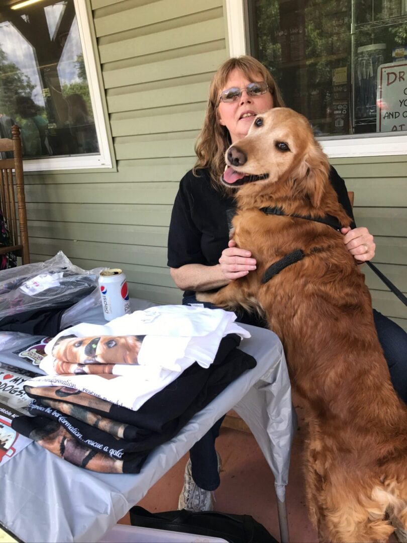 A woman sitting at a table with a golden retriever in her lap, merchandise on the table in front of them.