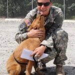 A soldier in camouflage uniform kneels and smiles while hugging a happy-looking dog outdoors.